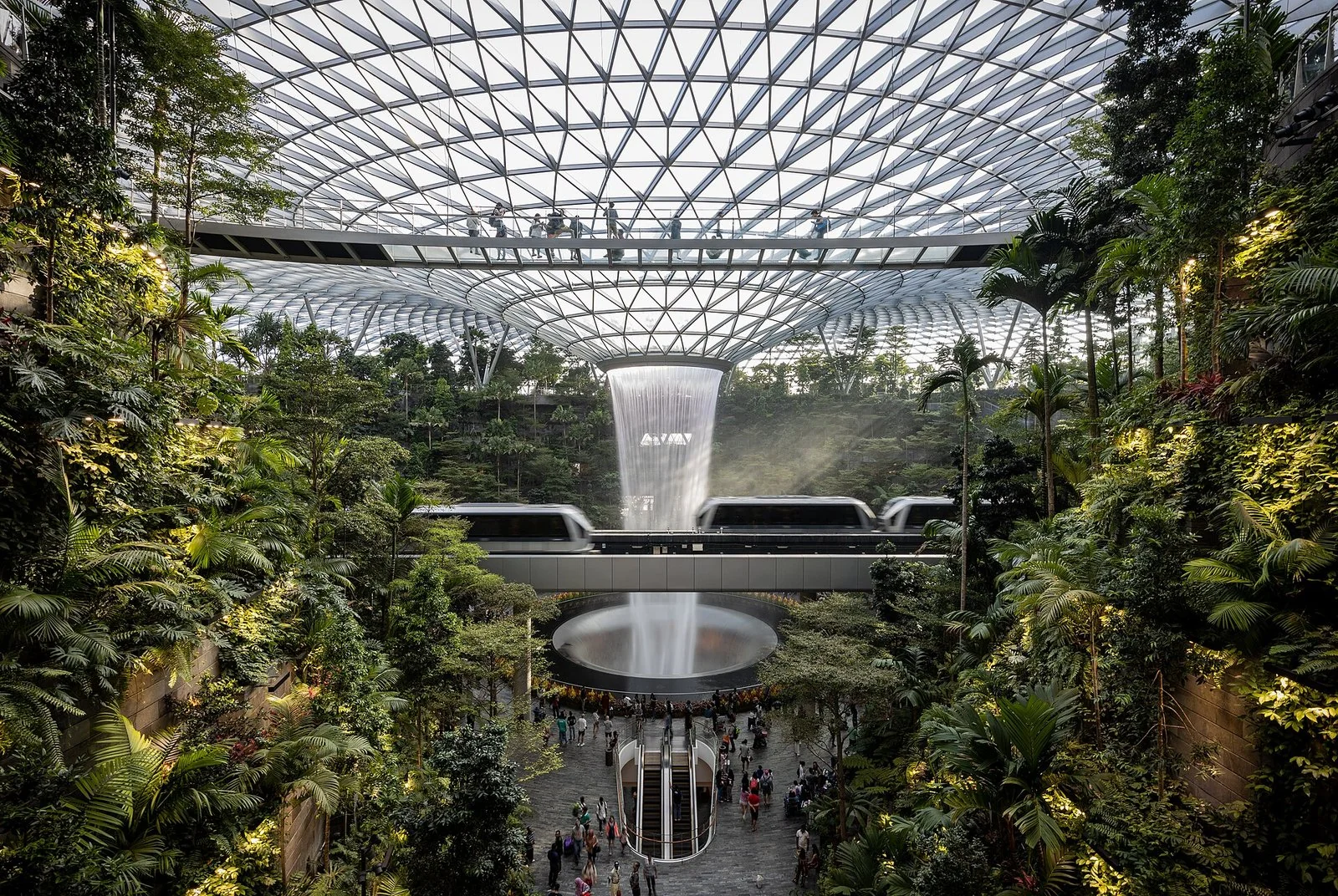 チャンギ空港ジュエルのガラスの屋根の下に落ちる屋内の滝と、木々の間を走る空港の電車の写真。