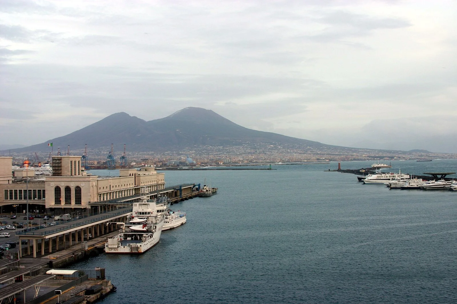 曇り空の下、ナポリの港の岸壁と海の向こうに、ふもとまで町が続くヴェスヴィオ山。