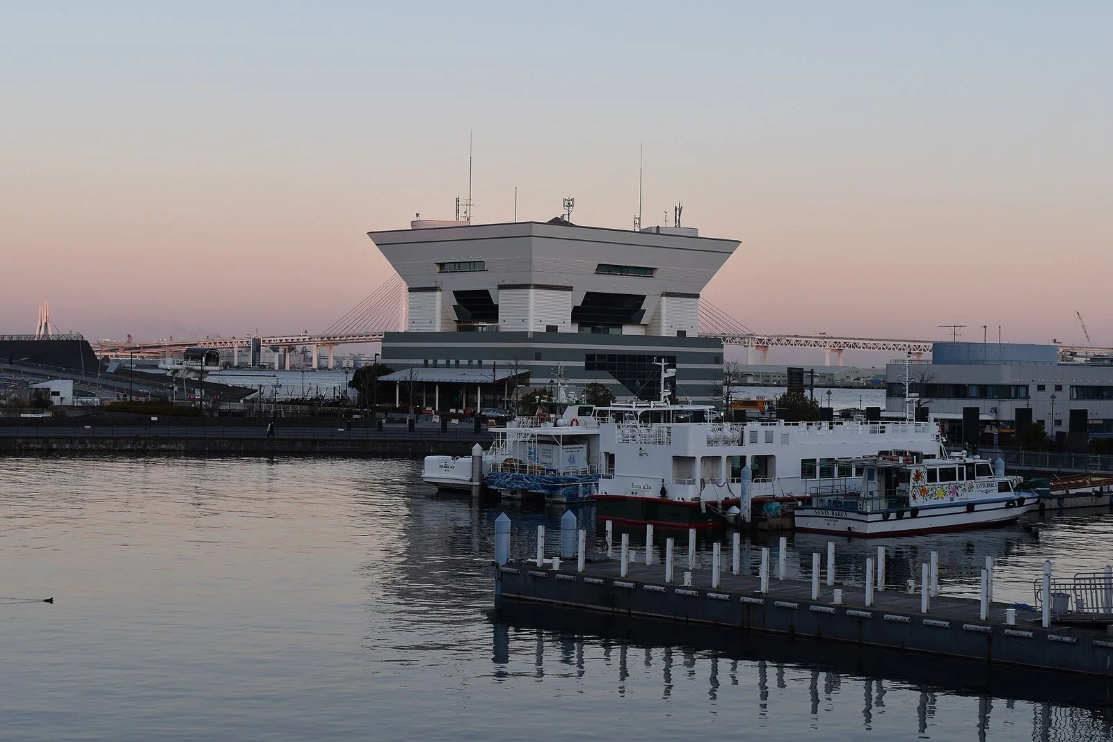 夕焼けの空の下、水辺に建つ平たい屋根のターミナル。手前に小さな船が泊まっている。