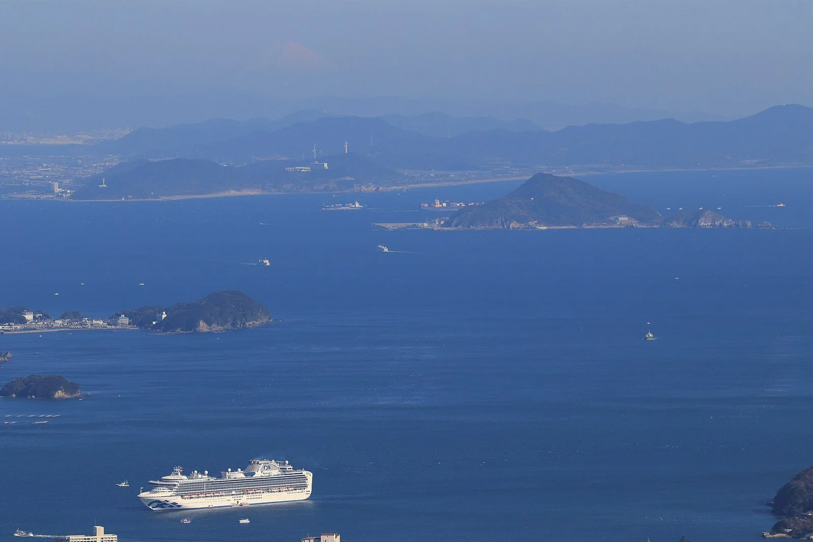青い海に白い大型客船が浮かび、奥に島と半島、遠くに富士山の頂がかすむ。