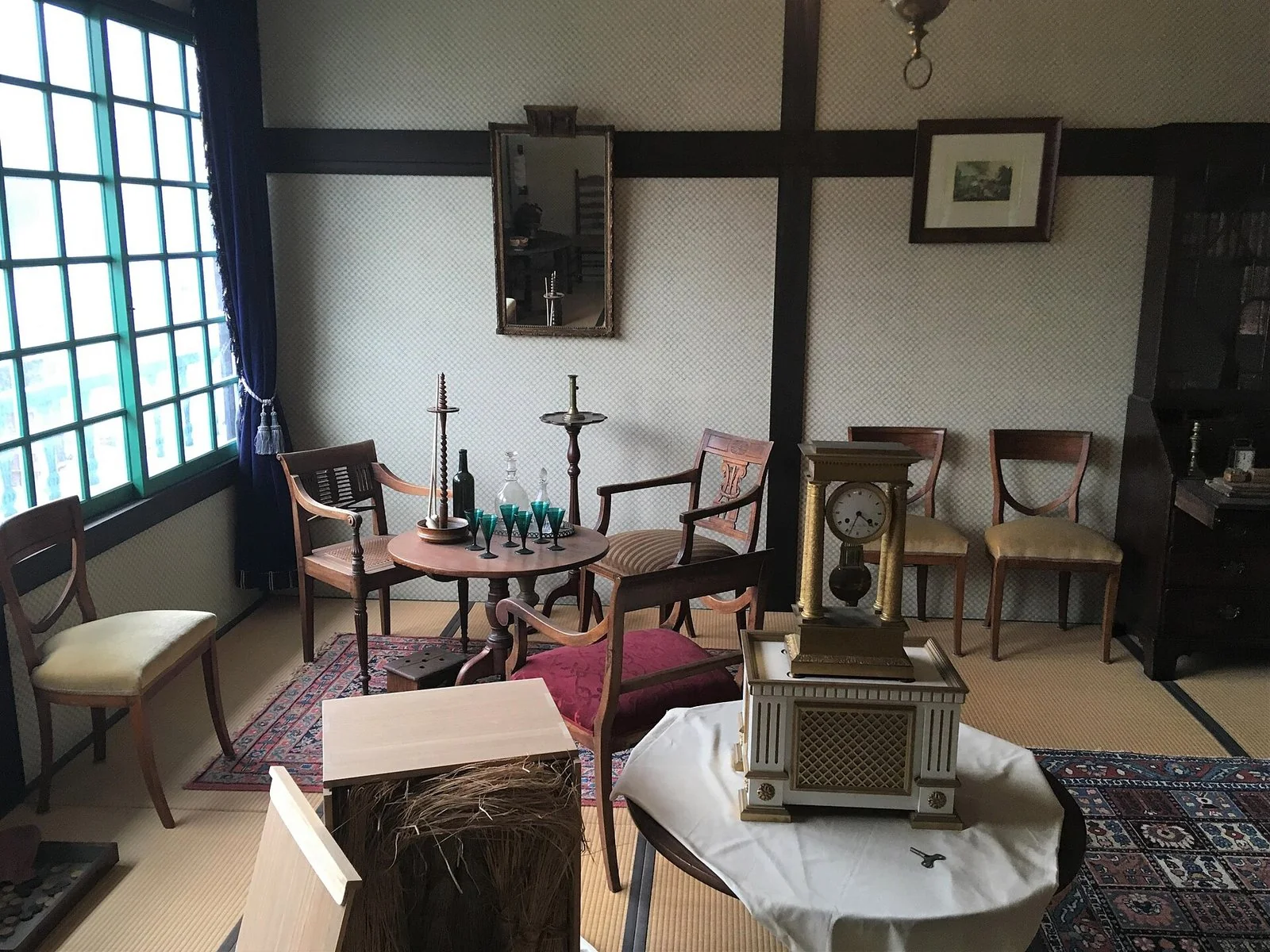 畳の部屋に、木の椅子と丸いテーブル、緑のガラスの杯、金色の置き時計が並ぶ。左に格子の窓がある。