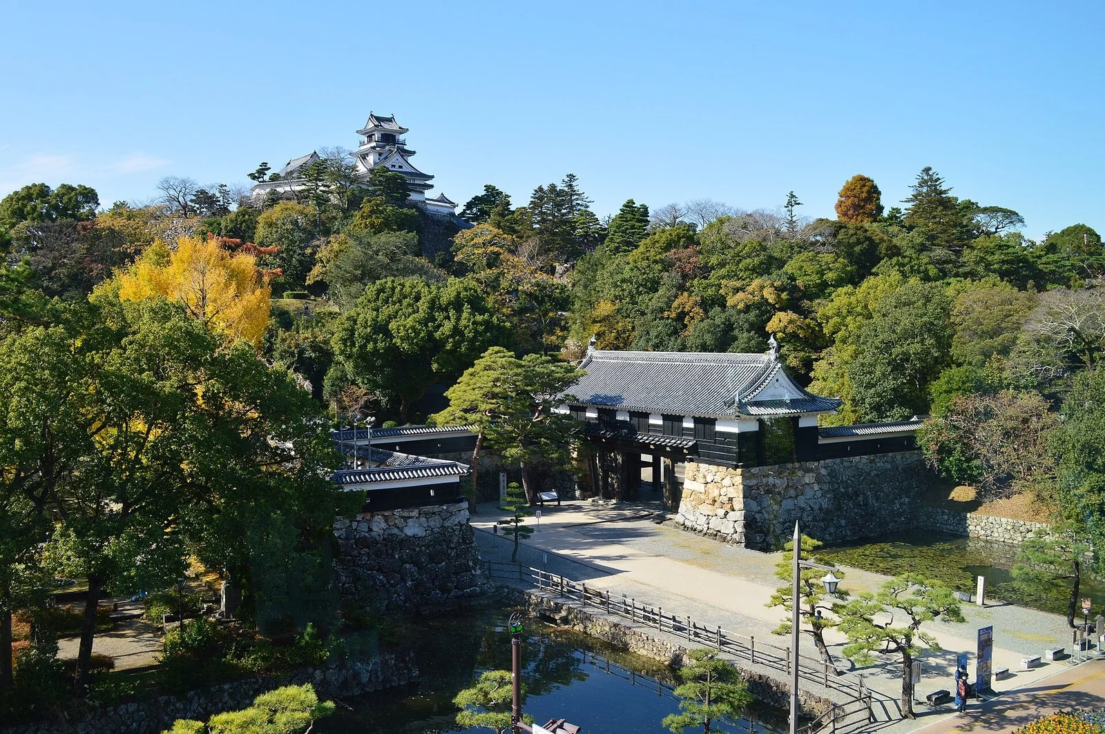 木々の茂る丘の上に白い天守が立ち、手前に堀と、石垣の上に建つ黒い門がある。
