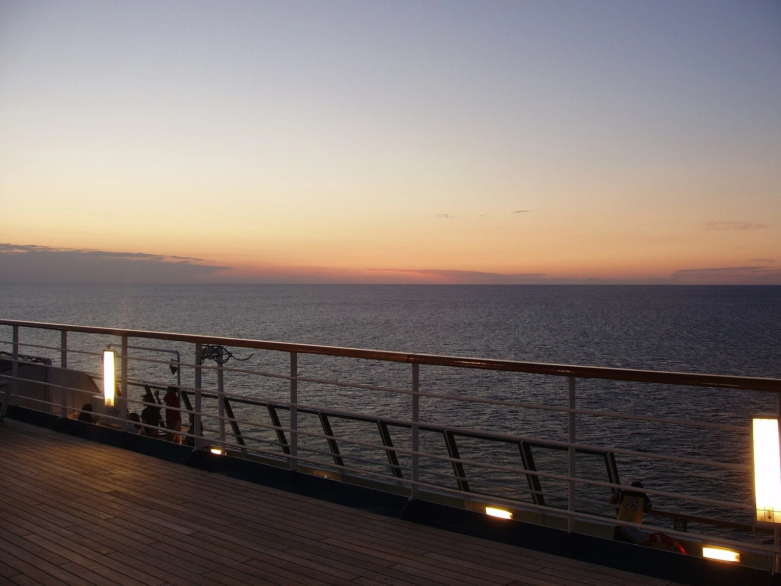木の甲板と白い手すりの向こうに、夕焼けの水平線と穏やかな海が広がる。手すりの灯りがともり、奥に小さな人影がある。